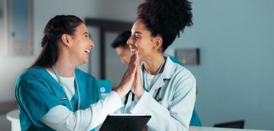 Duas profissionais de saúde sorrindo e se cumprimentando com as mãos em um ambiente de trabalho. A imagem transmite alegria e colaboração no setor de saúde.