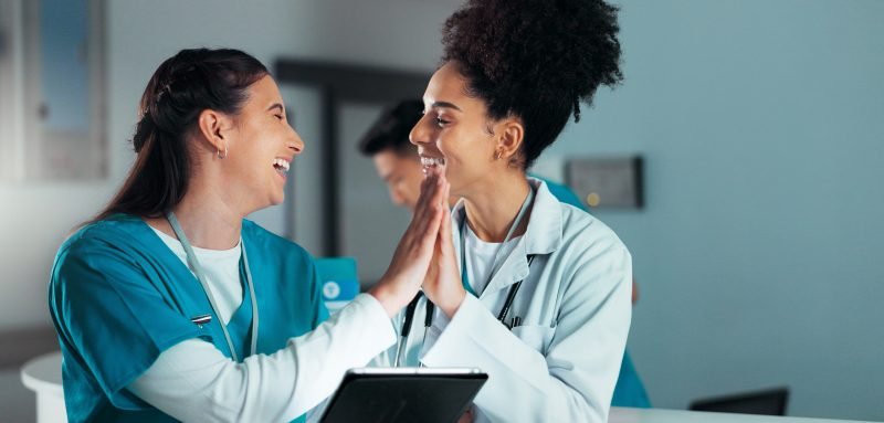 Duas profissionais de saúde sorrindo e se cumprimentando com as mãos em um ambiente de trabalho. A imagem transmite alegria e colaboração no setor de saúde.