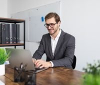 Homem sorridente trabalhando em um escritório moderno com laptop, ambientes organizados e plantas. Ideal para temas de produtividade e trabalho.