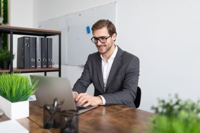 Homem sorridente trabalhando em um escritório moderno com laptop, ambientes organizados e plantas. Ideal para temas de produtividade e trabalho.