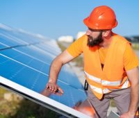 Técnico ajustando painel solar em um projeto de energia renovável, usando capacete laranja e colete refletivo, em campo aberto com céu azul.