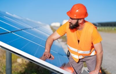Técnico ajustando painel solar em um projeto de energia renovável, usando capacete laranja e colete refletivo, em campo aberto com céu azul.