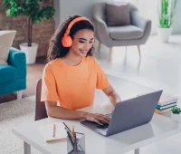 Jovem mulher sorridente usando fones de ouvido laranja enquanto trabalha em um laptop em um ambiente moderno e iluminado, ideal para teletrabalho.