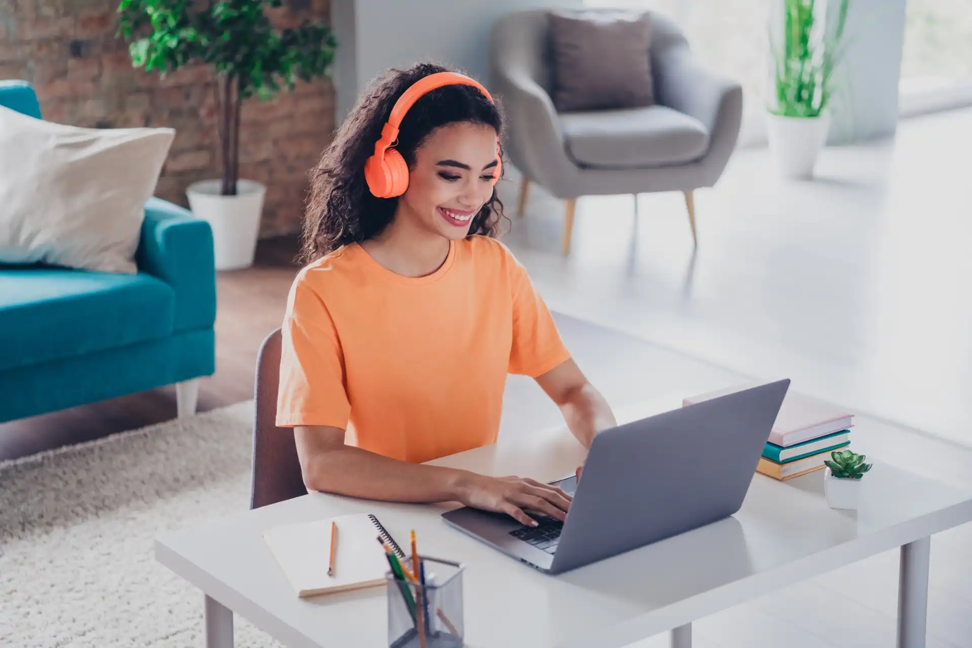 Jovem mulher sorridente usando fones de ouvido laranja enquanto trabalha em um laptop em um ambiente moderno e iluminado, ideal para teletrabalho.