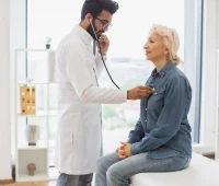 Um médico examina uma paciente sorridente em um consultório. O profissional utiliza um estetoscópio para avaliar a saúde da mulher, que parece tranquila e confiante.
