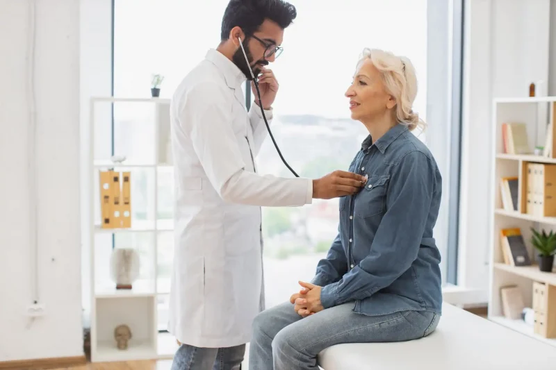Um médico examina uma paciente sorridente em um consultório. O profissional utiliza um estetoscópio para avaliar a saúde da mulher, que parece tranquila e confiante.