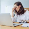 Mulher estressada com dores de cabeça enquanto trabalha em seu laptop em casa, com papéis e óculos na mesa. Acessórios de trabalho estão ao seu redor.