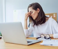 Mulher estressada com dores de cabeça enquanto trabalha em seu laptop em casa, com papéis e óculos na mesa. Acessórios de trabalho estão ao seu redor.