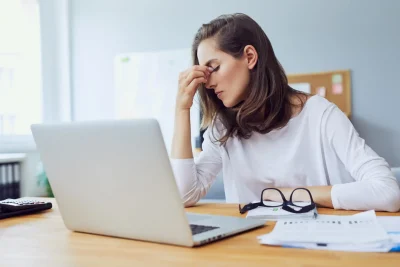 Mulher estressada com dores de cabeça enquanto trabalha em seu laptop em casa, com papéis e óculos na mesa. Acessórios de trabalho estão ao seu redor.