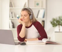Mulher utilizando fones de ouvido enquanto estuda em frente ao computador, com livro aberto e caneta na mão, em ambiente claro e moderno.