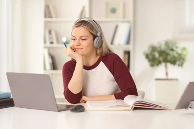 Mulher utilizando fones de ouvido enquanto estuda em frente ao computador, com livro aberto e caneta na mão, em ambiente claro e moderno.