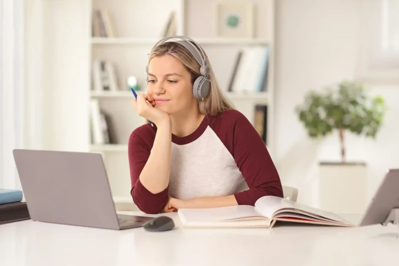 Mulher utilizando fones de ouvido enquanto estuda em frente ao computador, com livro aberto e caneta na mão, em ambiente claro e moderno.