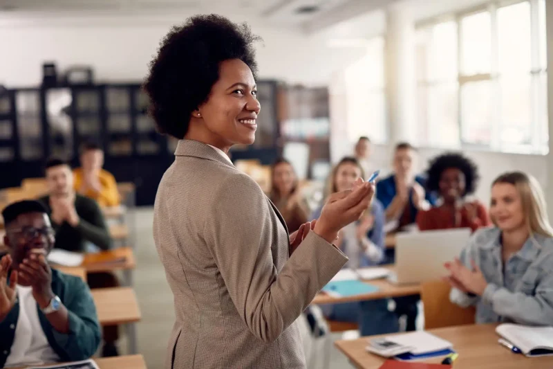 Professora sorridente interagindo com alunos em sala de aula, promovendo um ambiente de aprendizado inspirador e motivador. Alunos aplaudindo, destacando a importância da educação.