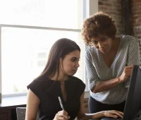 Imagem de duas mulheres colaborando em um ambiente de escritório, uma ajudando a outra com o uso de um computador.
