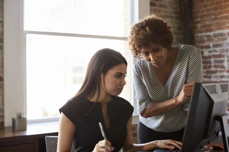 Imagem de duas mulheres colaborando em um ambiente de escritório, uma ajudando a outra com o uso de um computador.