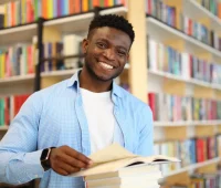 Jovem sorridente em biblioteca segurando um livro em frente a uma estante cheia de livros coloridos, representando o amor pela leitura e educação.