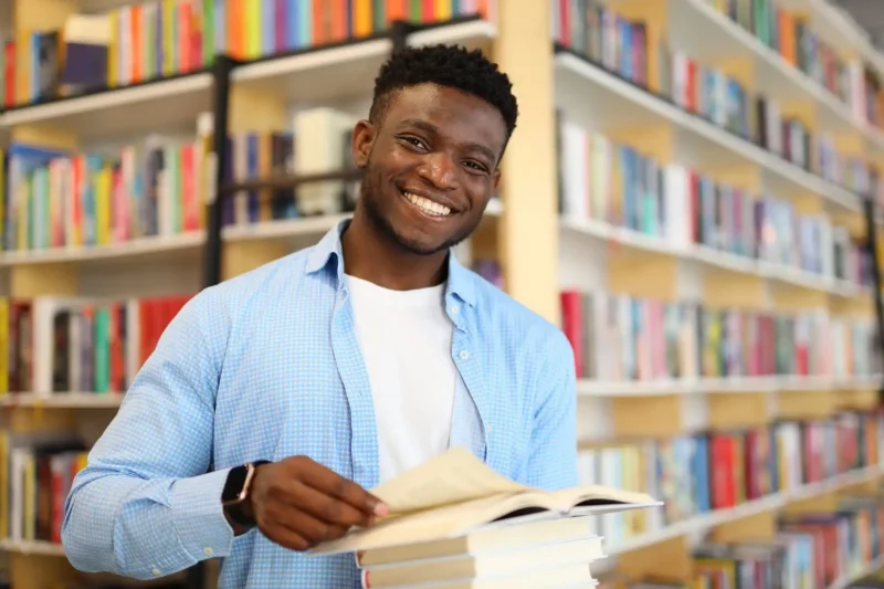 Jovem sorridente em biblioteca segurando um livro em frente a uma estante cheia de livros coloridos, representando o amor pela leitura e educação.