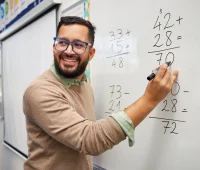 Professor sorridente explicando operações matemáticas no quadro branco, ensinando adição e subtração para alunos em sala de aula.