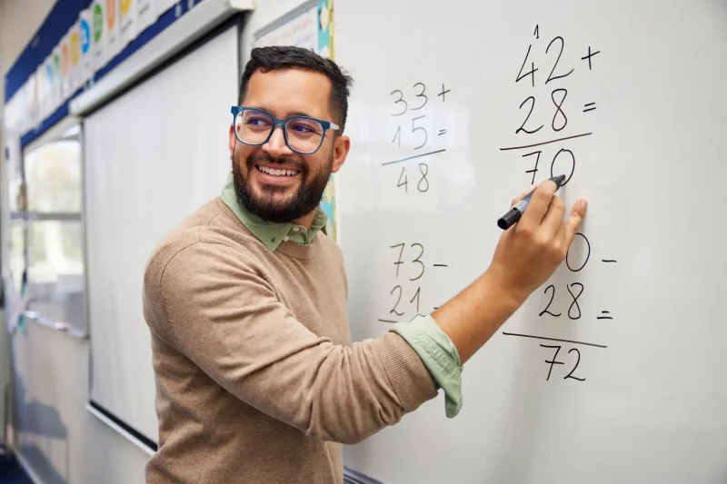 Professor sorridente explicando operações matemáticas no quadro branco, ensinando adição e subtração para alunos em sala de aula.