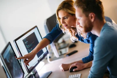 Profissionais colaborando em um ambiente de trabalho moderno, analisando dados em computadores. A imagem retrata a dinâmica de trabalho em equipe na tecnologia.