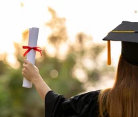 Estudante graduada segurando um diploma com fita vermelha, simbolizando a conquista da formatura, ao fundo um ambiente natural iluminado.