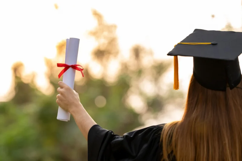 Estudante graduada segurando um diploma com fita vermelha, simbolizando a conquista da formatura, ao fundo um ambiente natural iluminado.
