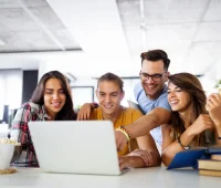 Grupo de jovens se divertindo enquanto assistem a um vídeo em um laptop. Eles demonstram alegria e camaradagem em um ambiente moderno.