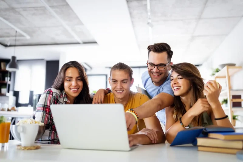 Grupo de jovens se divertindo enquanto assistem a um vídeo em um laptop. Eles demonstram alegria e camaradagem em um ambiente moderno.