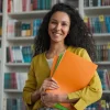 Mulher sorridente em biblioteca, segurando pastas coloridas, simbolizando organização e estudos, ideal para conteúdo educacional.