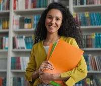 Mulher sorridente em biblioteca, segurando pastas coloridas, simbolizando organização e estudos, ideal para conteúdo educacional.