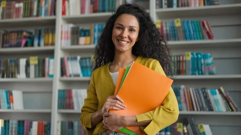 Mulher sorridente em biblioteca, segurando pastas coloridas, simbolizando organização e estudos, ideal para conteúdo educacional.