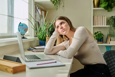 Mulher pensativa em ambiente de trabalho, refletindo sobre suas atividades diante do computador, com itens de escritório ao fundo.