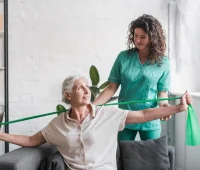 Mulher idosa realizando exercícios de fisioterapia com auxílio de uma profissional de saúde, usando faixa elástica para melhorar a mobilidade e força.