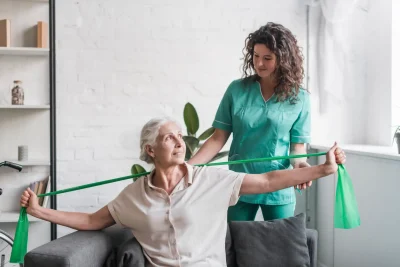Mulher idosa realizando exercícios de fisioterapia com auxílio de uma profissional de saúde, usando faixa elástica para melhorar a mobilidade e força.