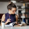 Jovem leitor estudando em biblioteca, segurando uma caneta e livros, demonstrando concentração e dedicação ao aprendizado.