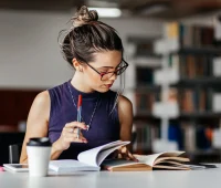 Jovem leitor estudando em biblioteca, segurando uma caneta e livros, demonstrando concentração e dedicação ao aprendizado.