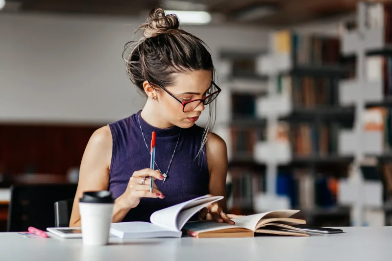 Jovem leitor estudando em biblioteca, segurando uma caneta e livros, demonstrando concentração e dedicação ao aprendizado.