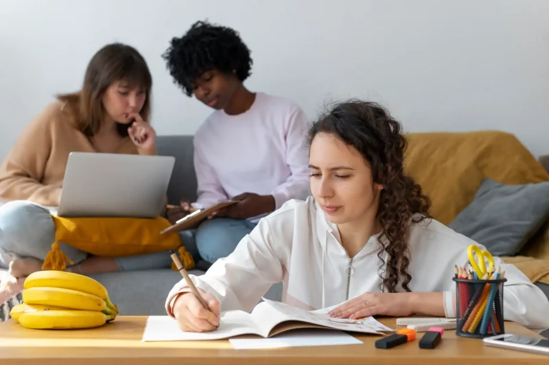 Três estudantes se preparam revisando os assuntos que mais caem no Enem, com livros, laptop e materiais de estudo espalhados, em ambiente confortável e colaborativo.