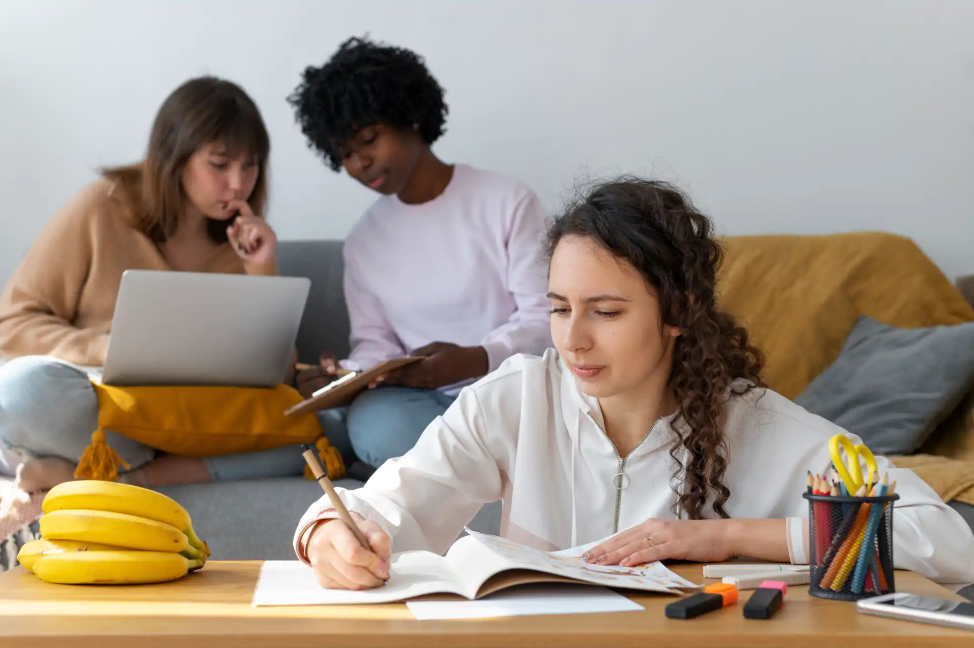 Três estudantes se preparam revisando os assuntos que mais caem no Enem, com livros, laptop e materiais de estudo espalhados, em ambiente confortável e colaborativo.