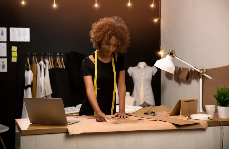 Mulher estilista no ateliê de moda, medindo e ajustando tecido de papelão para confecção de roupas, com prancheta, laptop e plantas decorativas ao fundo.