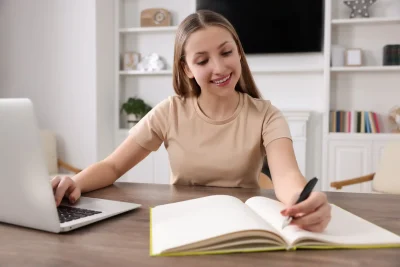 Jovem mulher estudando com caderno aberto e livro em uma mesa, usando laptop em um ambiente bem iluminado e organizado