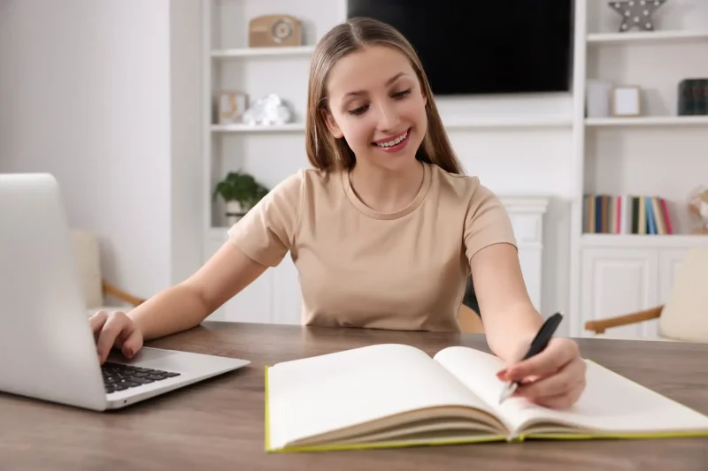 Jovem mulher estudando com caderno aberto e livro em uma mesa, usando laptop em um ambiente bem iluminado e organizado