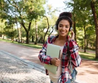 Jovem estudante sorridente usando fones de ouvido e carregando um caderno enquanto caminha por um parque ao ar livre.