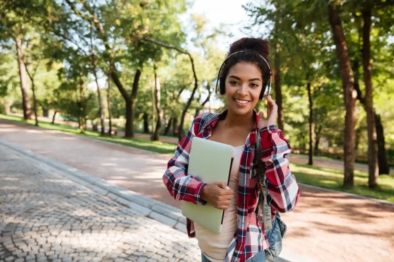 Jovem estudante sorridente usando fones de ouvido e carregando um caderno enquanto caminha por um parque ao ar livre.