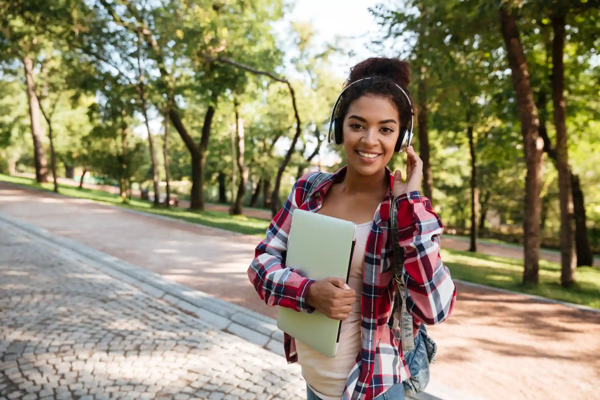 Jovem estudante sorridente usando fones de ouvido e carregando um caderno enquanto caminha por um parque ao ar livre.