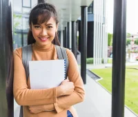 Jovem estudante sorridente segurando caderno, vestindo moletom bege e mochila, ao lado de prédio moderno na rua, representando sucesso estudantil e educação.