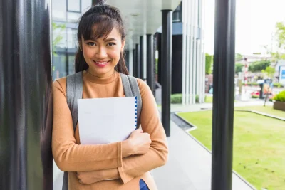 Jovem estudante sorridente segurando caderno, vestindo moletom bege e mochila, ao lado de prédio moderno na rua, representando sucesso estudantil e educação.
