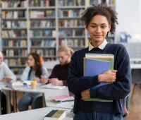 Jovem estudante sorridente segurando cadernos e livros na biblioteca, em ambiente de aprendizado com outros profissionais ao fundo.