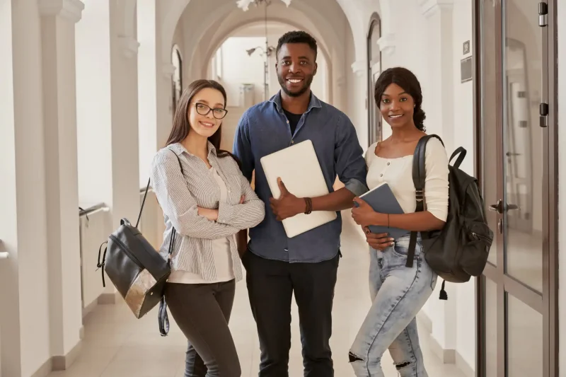 Três estudantes universitários sorridentes posando juntos no corredor de uma faculdade, carregando laptops e mochilas, representando sucesso acadêmico e amizade.
