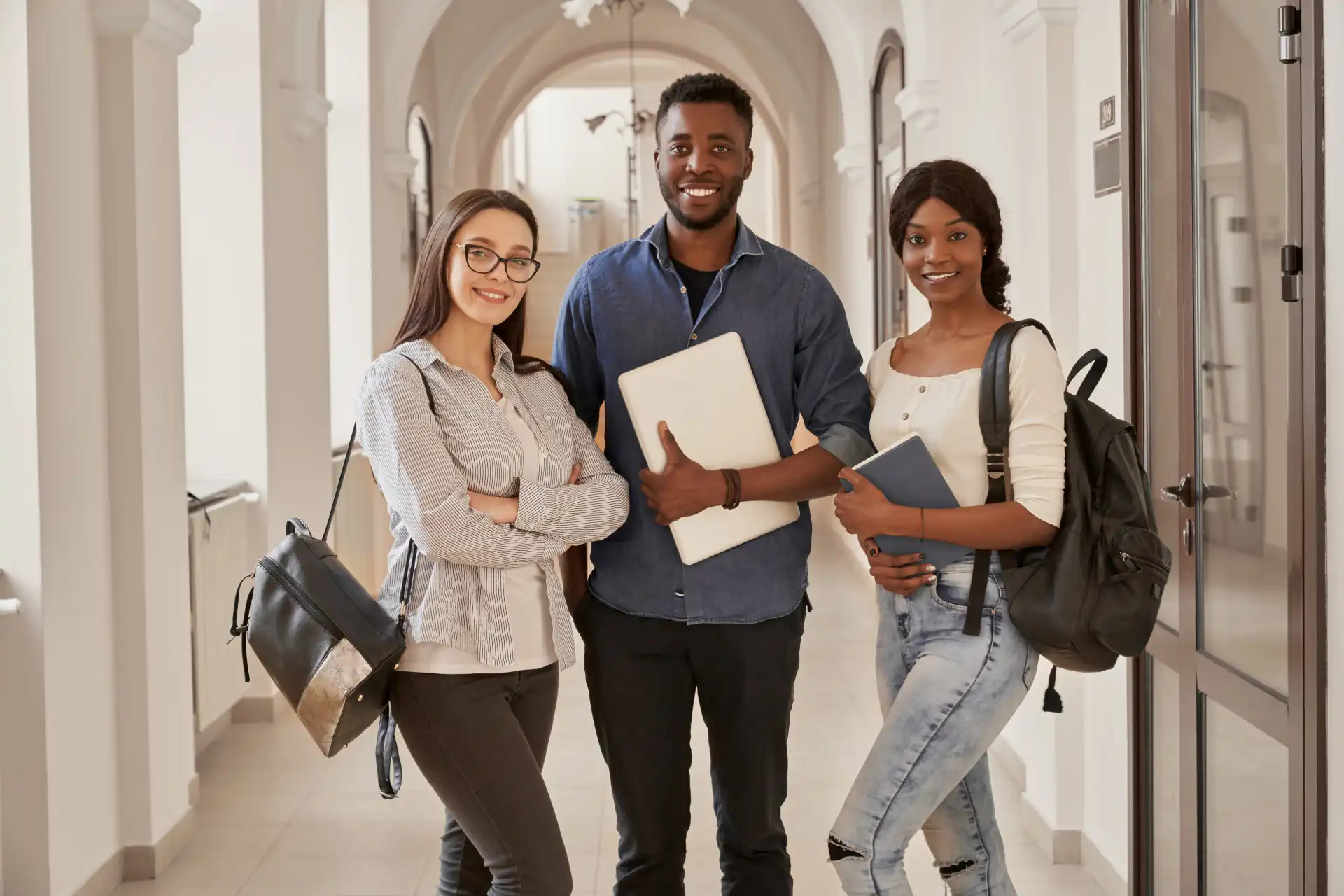 Três estudantes universitários sorridentes posando juntos no corredor de uma faculdade, carregando laptops e mochilas, representando sucesso acadêmico e amizade.